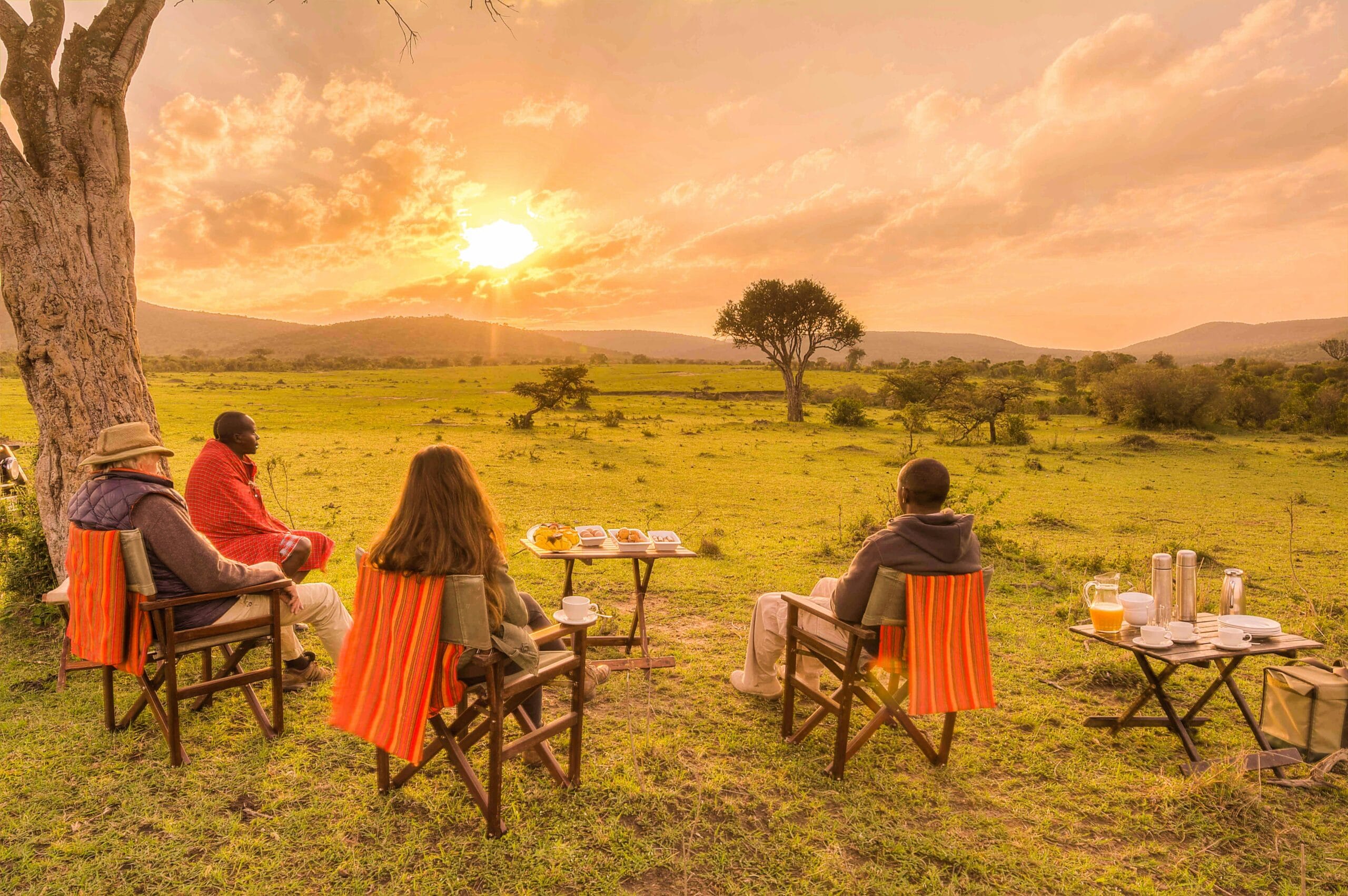 Four people enjoy a sunset picnic in the African savanna, surrounded by nature and serene views.